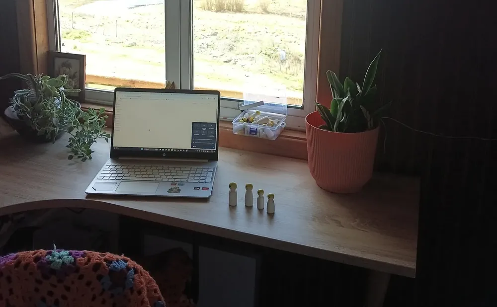 Escritorio de Maritza junto a la ventana, con un computador, plantas y cuatro pequeñas figuras de madera; afuera, la estepa